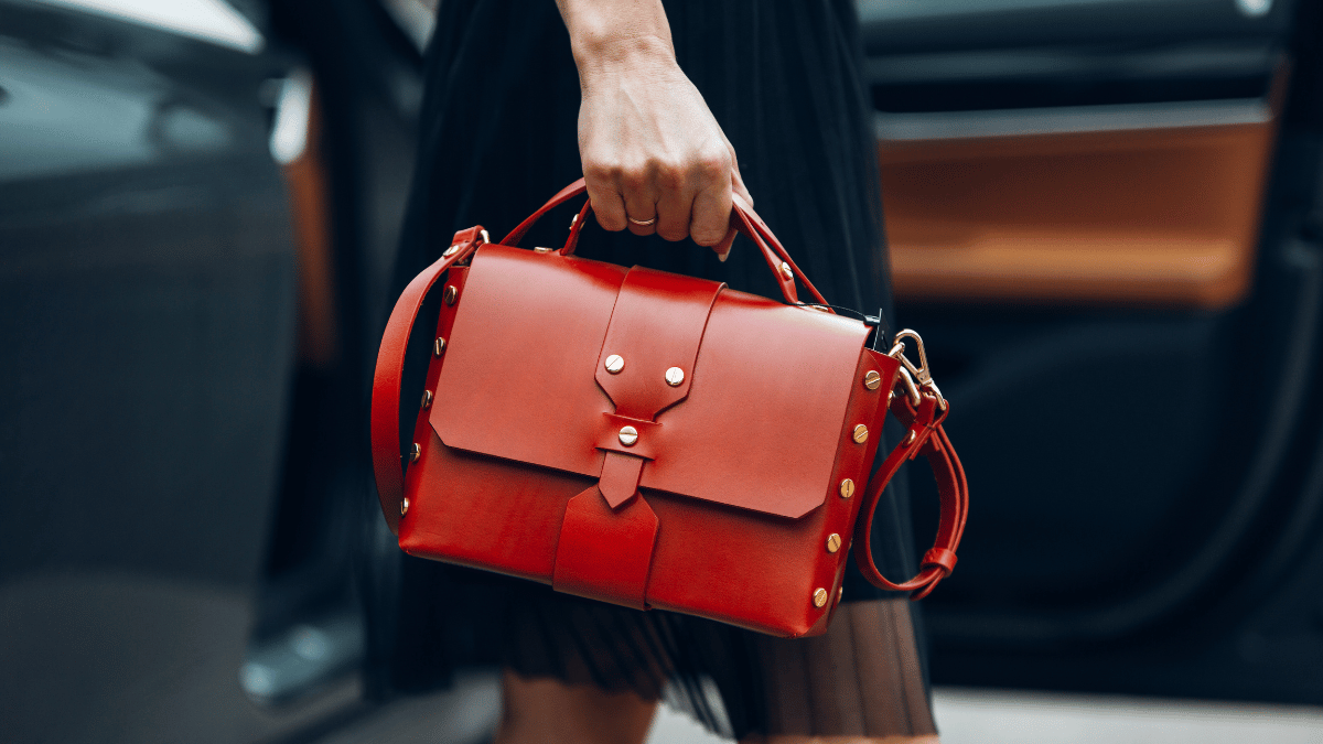 Imagem de uma mulher (apenas mostrando mão, parte da cintura e parte das pernas) segurando uma bolsa na lateral do corpo. A mulher está usando um vestido preto e a bolsa é vermelha.