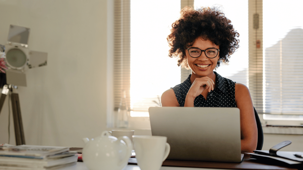 Imagem de uma mulher negra de pele clara com um black power. Ela está sentada à mesa em um escritório caseiro. Está sorrindo para a foto, com a mão no queixo e um notebook na sua frente.