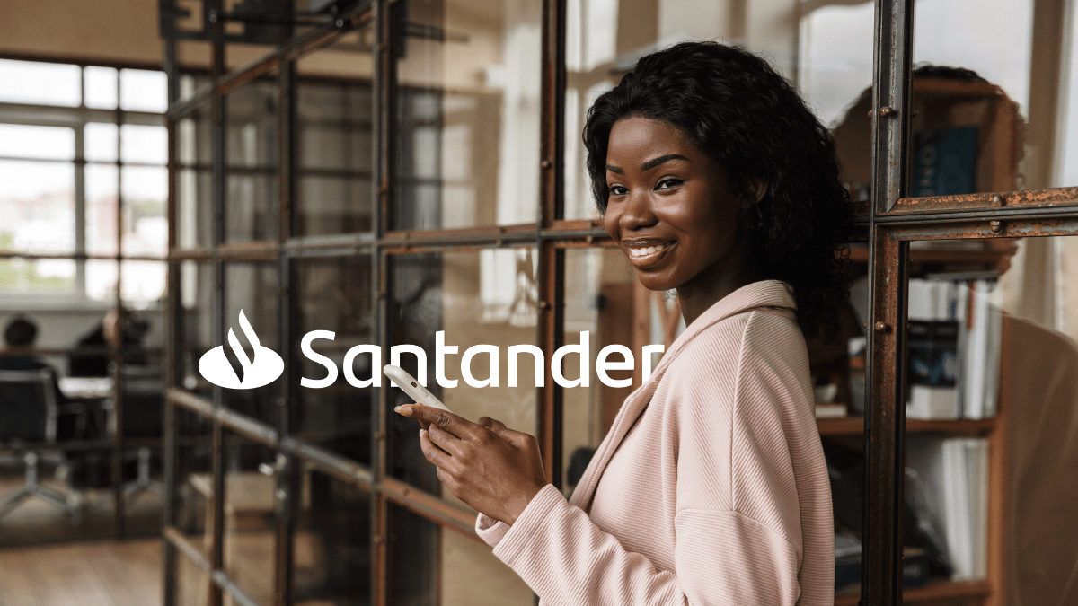 imagem de uma mulher negra de aproximadamente 30 anos. Ela está sorrindo, usando terno rosa claro em um ambiente externo a um escritório enquanto segura um celular. Ao fundo e lado dela está o logo do Santander.